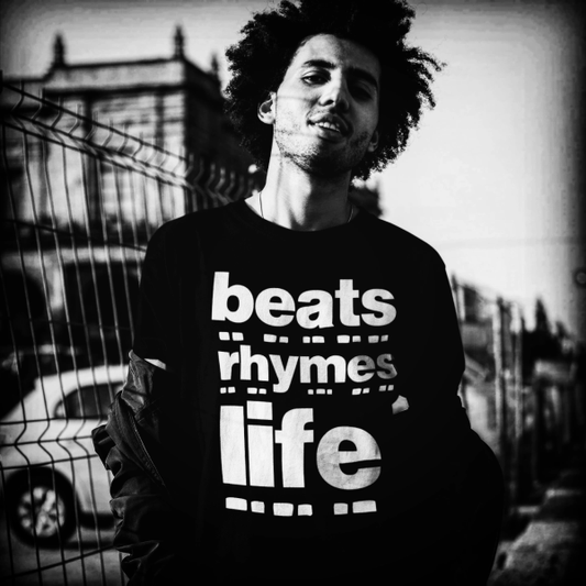 Young man with curly hair wearing a black long-sleeve T-shirt with bold white text reading “beats rhymes life,” standing outdoors by a wire fence in an urban setting, captured in high-contrast black and white.